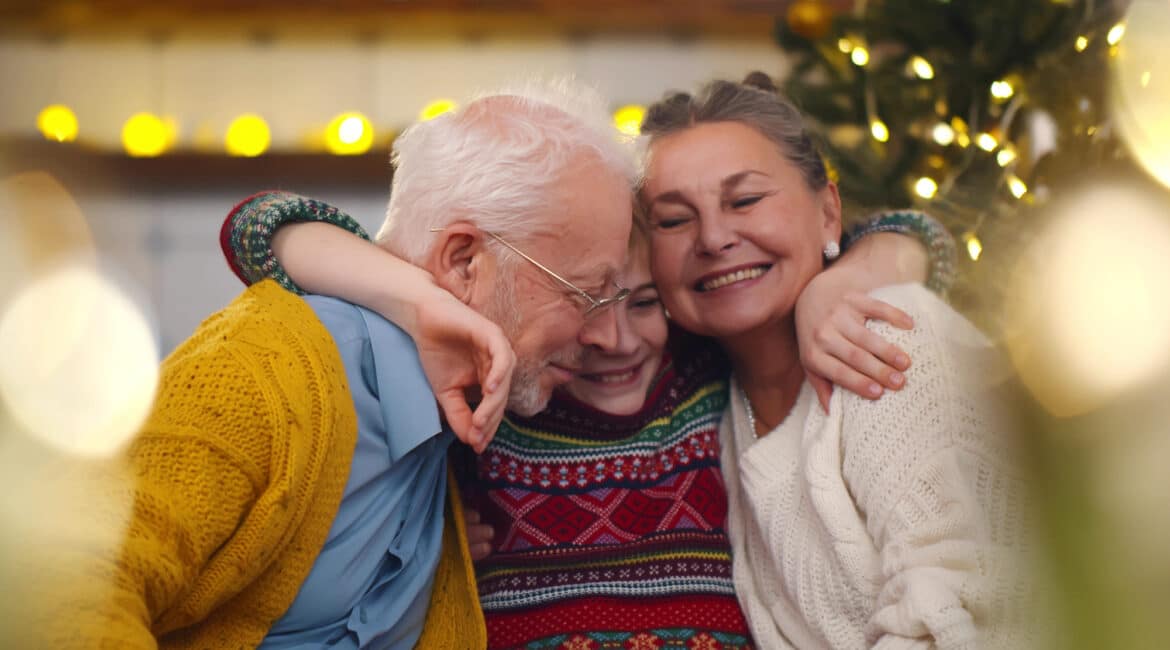 Smiling grandparents and grandson sitting on couch at home. Portrait of loving grandfather and grandmother embracing smiling preteen boy celebrating christmas together